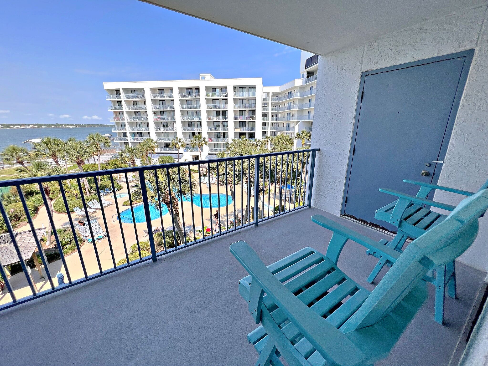 A balcony with turquoise chairs, a pool, and a view of a white building and beach.