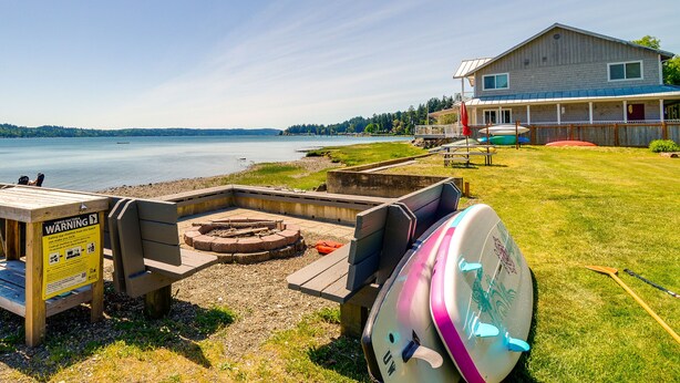 A lakeside property with a wooden deck, a covered seating area, and a large house with a porch. - Waterfront Hood Canal Cottage: Steps to the Beach! (Belfair)