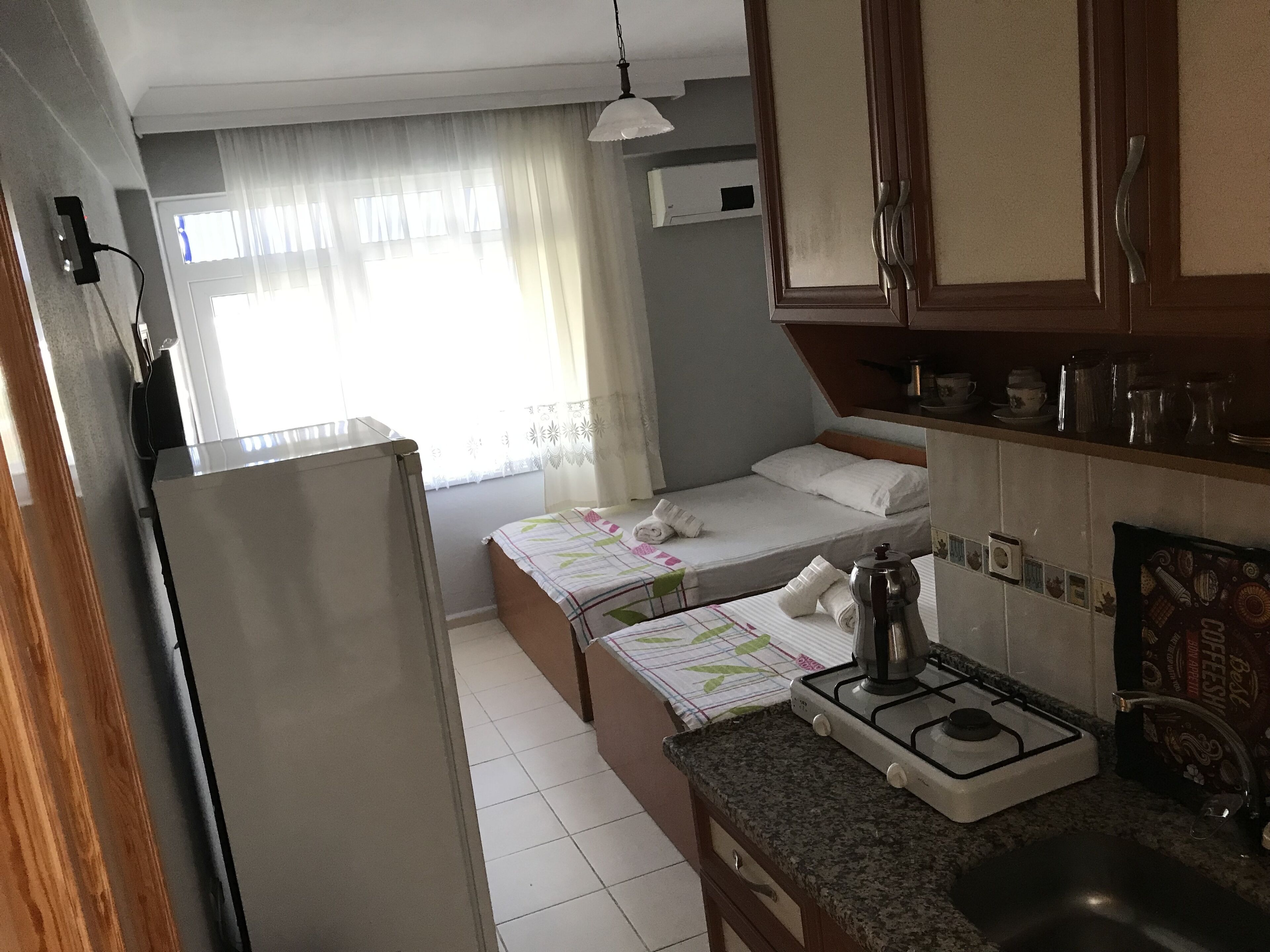Habitación de hotel compacta con kitchenette, cama con colcha de colores, ventana con cortinas translúcidas y un pequeño balcón.