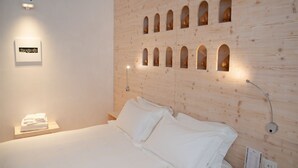 Quarto de hotel com cabeceira de madeira com vários recortes em arco, cama bem arrumada com roupa de cama branca e uma pequena mesa de cabeceira com abajur.
