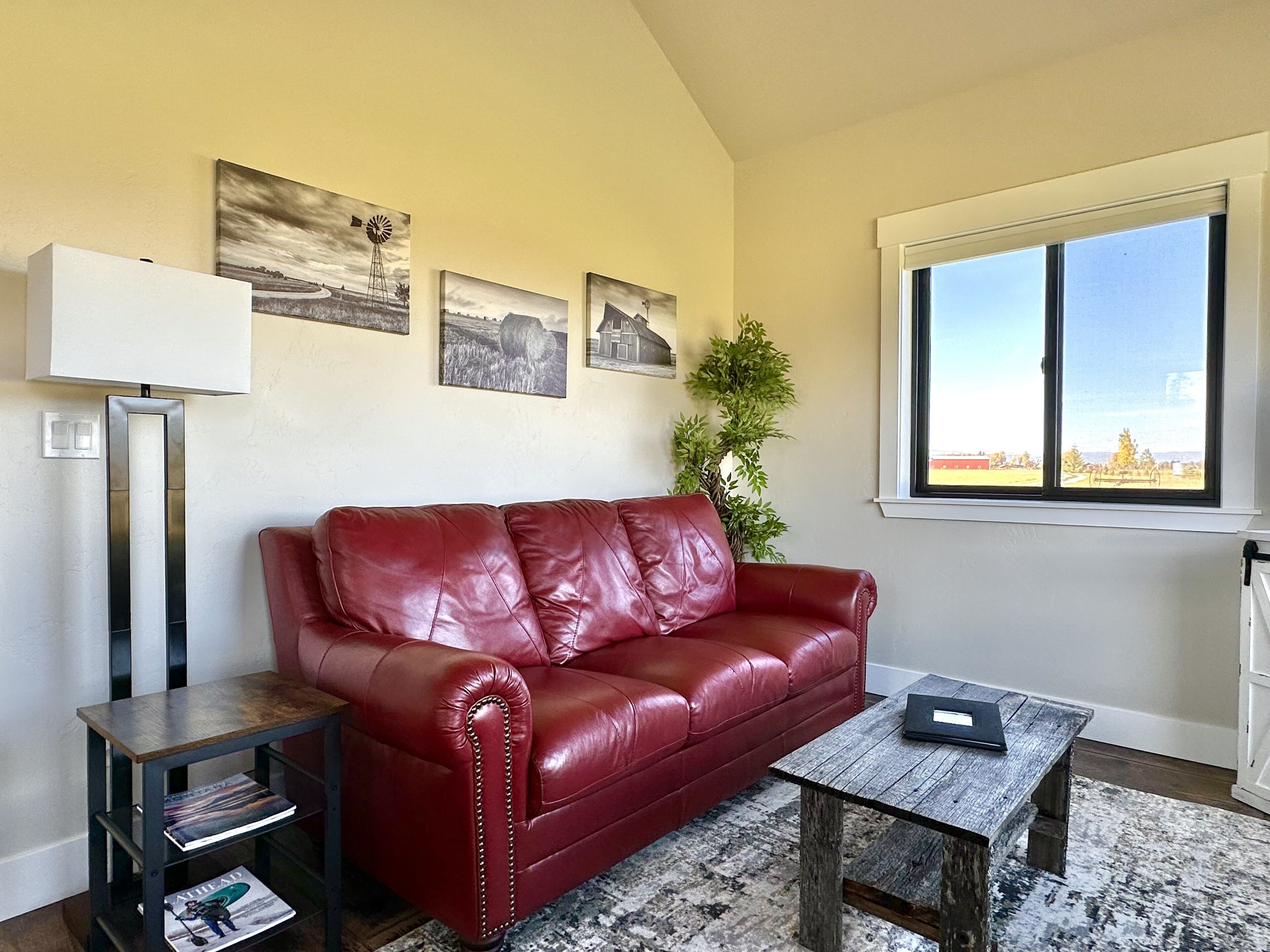 A living room with a red leather sofa, a wooden coffee table, a floor lamp, and a window with a view of a farm.