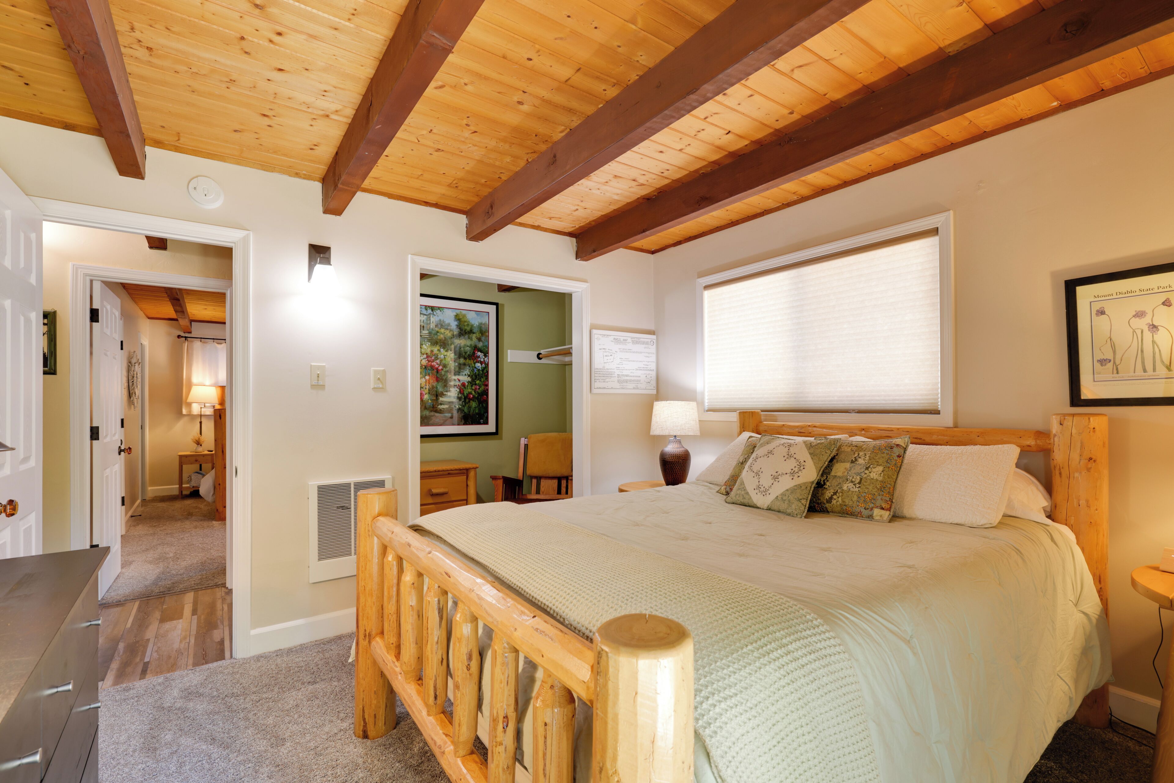 A bedroom with a wooden bed frame, a large window with blinds, and a painting on the wall.