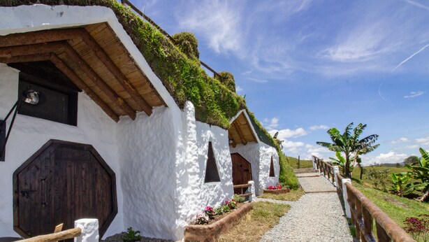 Un camino conduce a una hilera de casas blancas con techos de paja, puertas y ventanas de madera, rodeadas de vegetación y un cielo despejado.