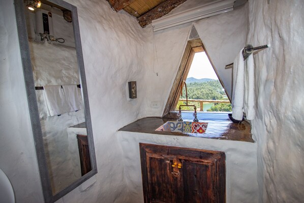 A rustic bathroom with a wooden cabinet, a sink with a colorful tile backsplash, and a view of a mountainous landscape through a triangular window. - HOBBIT HOTEL (El Peñol)