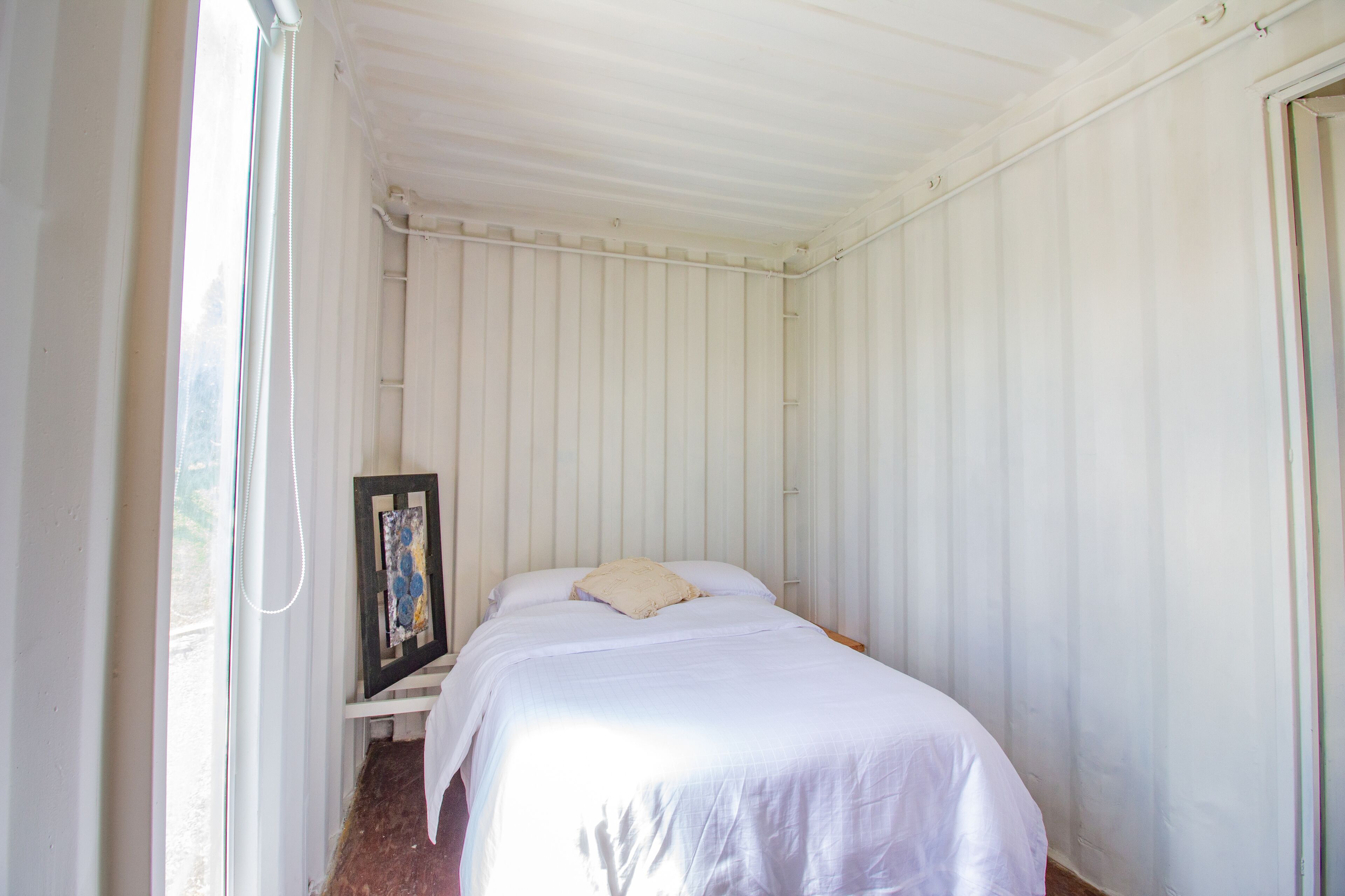 Habitació petita, de color blanc, amb un llit individual, una finestra a un costat i un quadre encimbellat a la paret.