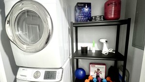 Un área de lavandería con lavadoras y secadoras apiladas, un estante con diversos artículos, incluyendo un extintor, una botella de agua y equipo de ejercicio.