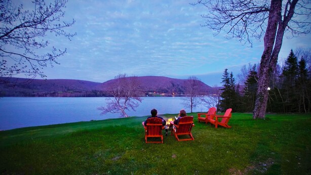 Two people sitting on red Adirondack chairs facing a lake with a hill in the background.