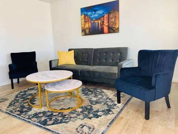 Una sala de estar con un sofá gris, un sillón azul, una mesa de centro redonda con tapa de mármol y una alfombra estampada.