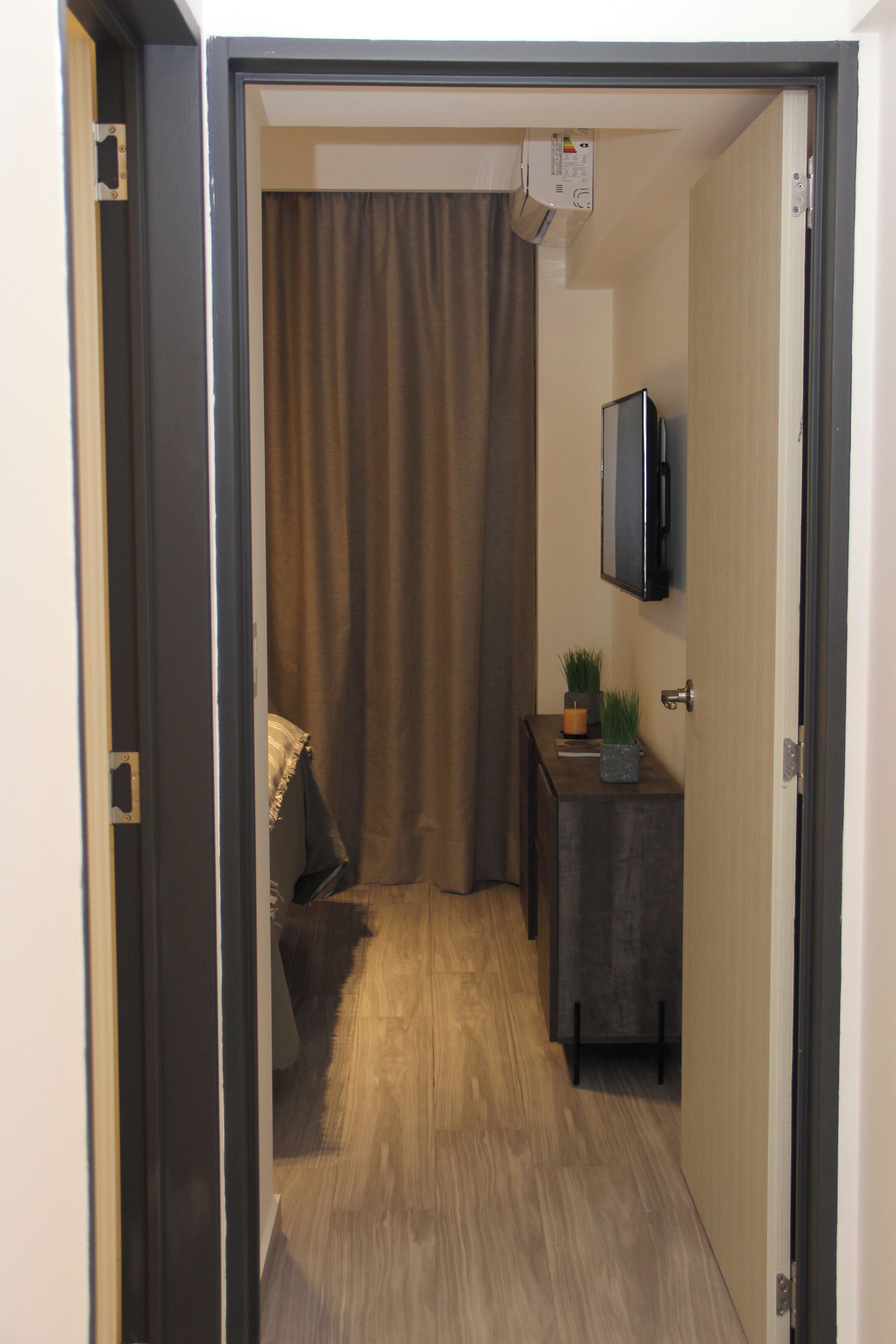 A narrow hallway with a wooden floor, a dark-colored door on the right, and a small table with a plant and a candle.