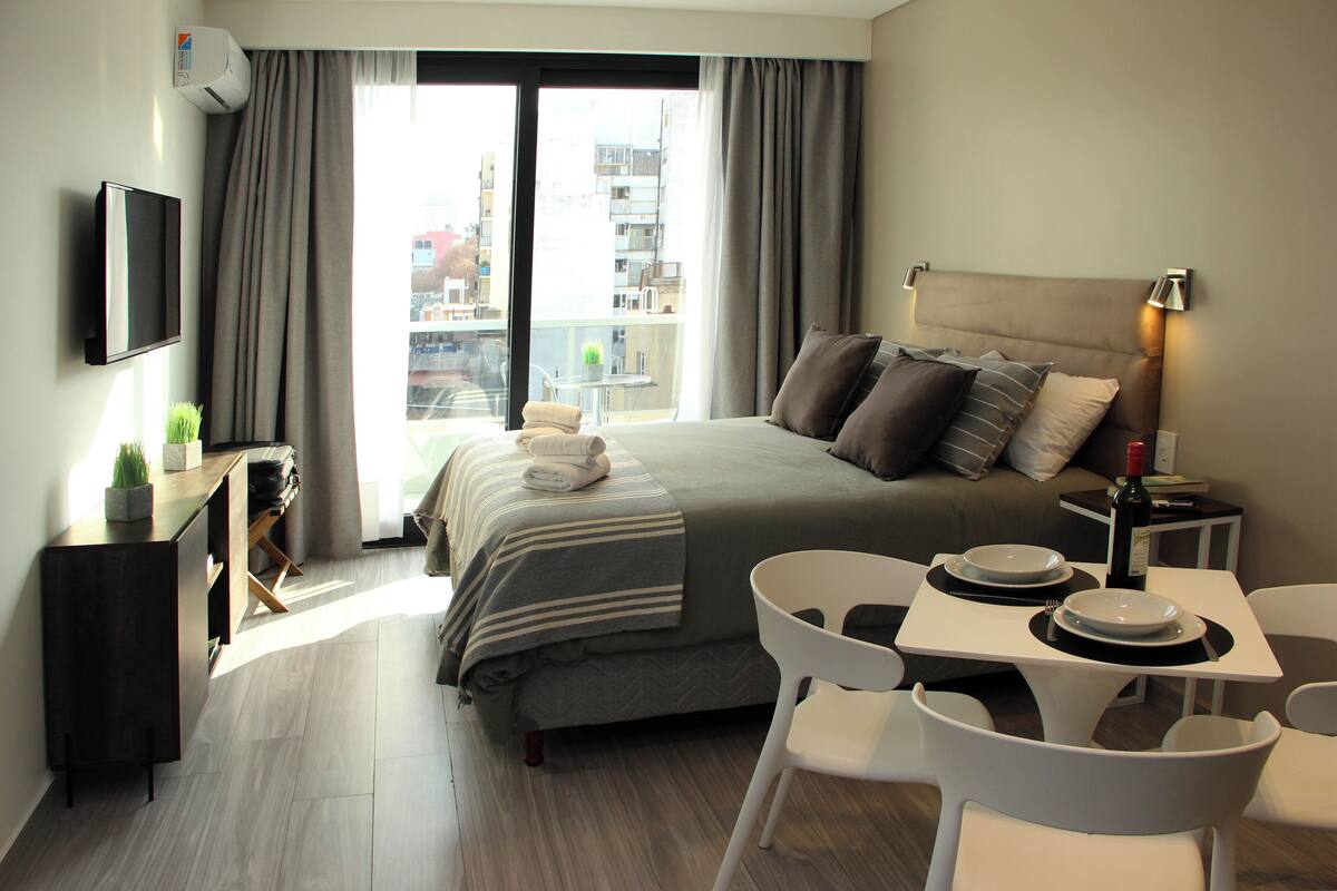 Un hotel moderno con una cama grande, una mesa de comedor con sillas, un televisor montado en la pared y un balcón con vistas a la ciudad.