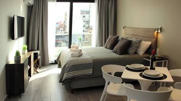 A modern hotel room with a large bed, a dining table with chairs, a TV mounted on the wall, and a balcony with a view of the city.