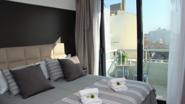 A hotel room with a large bed, two pillows, a bedside table with a lamp, and a view of the city through the window. - In House BA - Le Coq (Buenos Aires)
