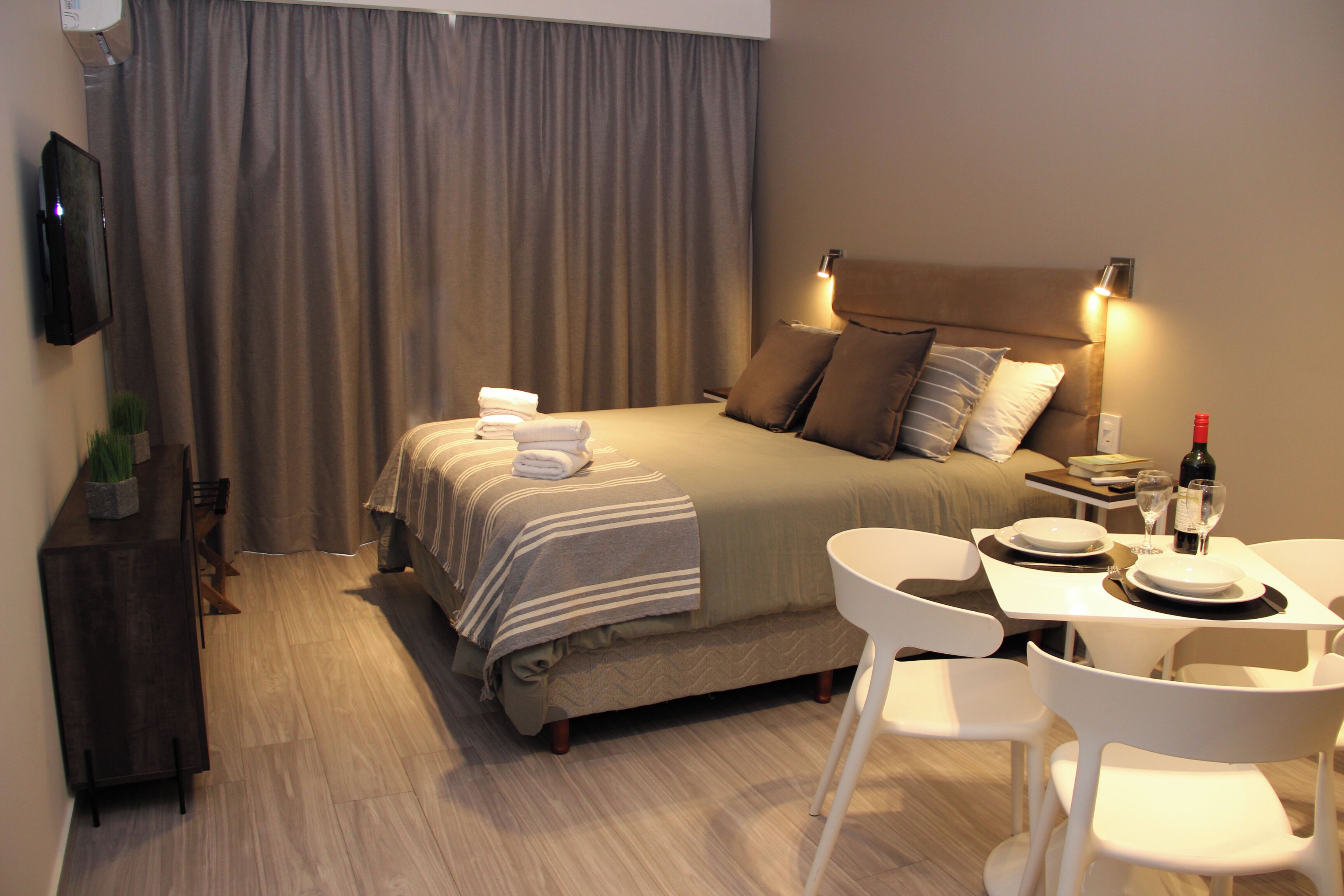 Quarto de hotel com cama, mesa de cabeceira, luminária e área de jantar com cadeiras e uma mesa.