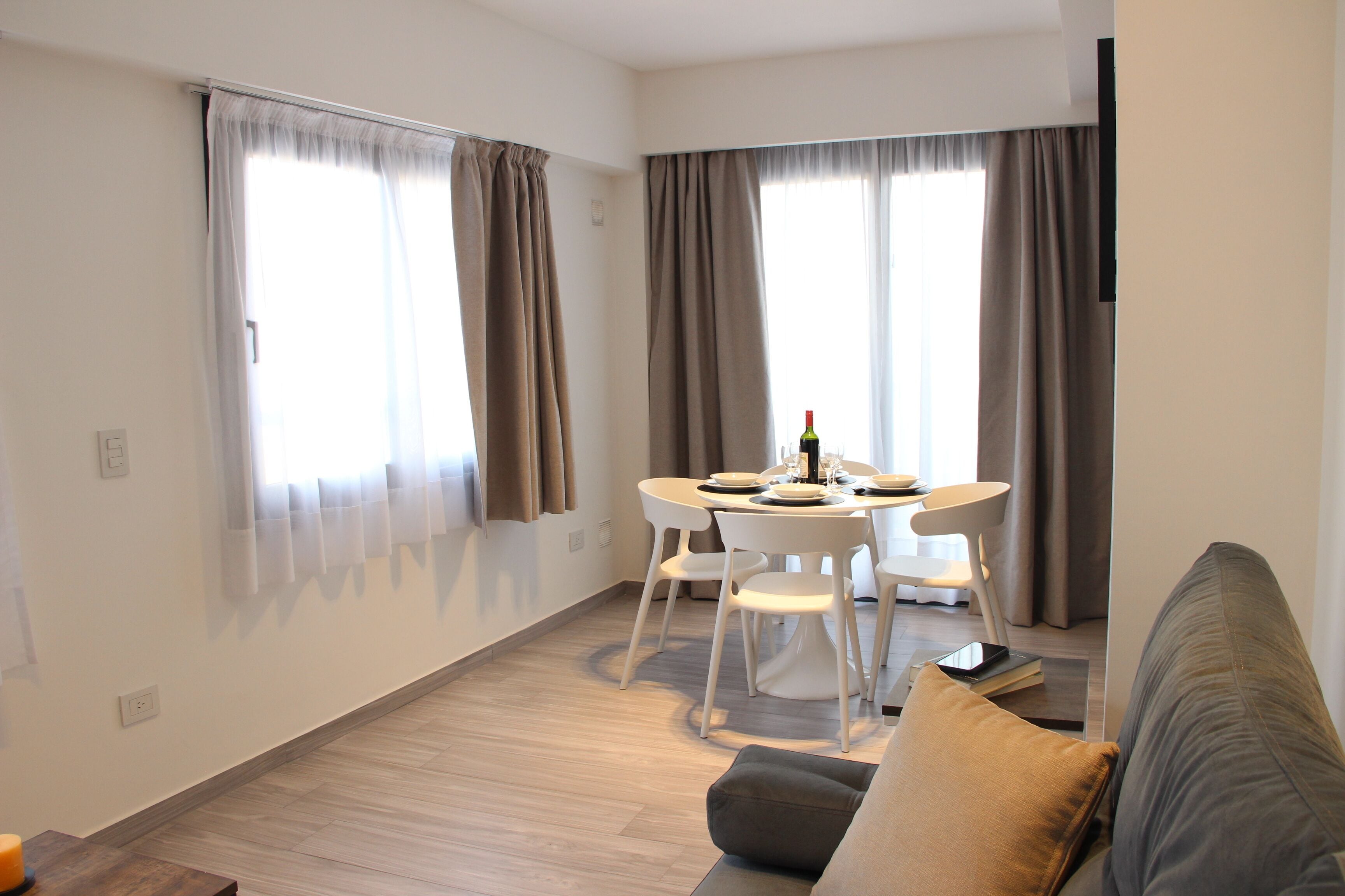 A modern living room with a dining area, featuring a sofa, a dining table with chairs, and large windows with curtains.