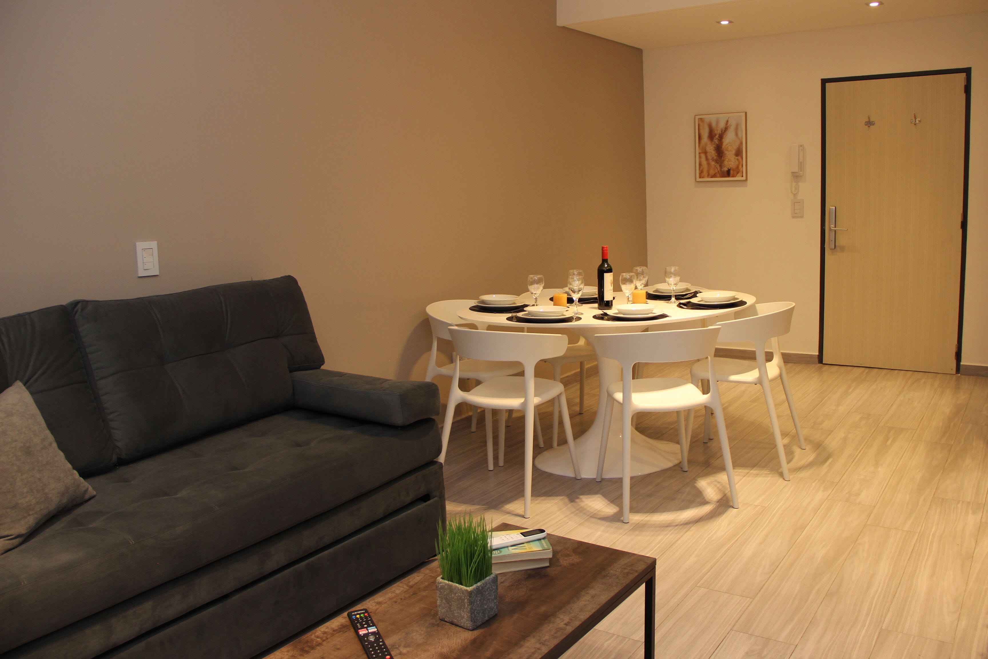 Una sala de estar moderna con un sofá gris, una mesa de comedor redonda con sillas blancas y una mesita de centro con un control remoto y una planta.