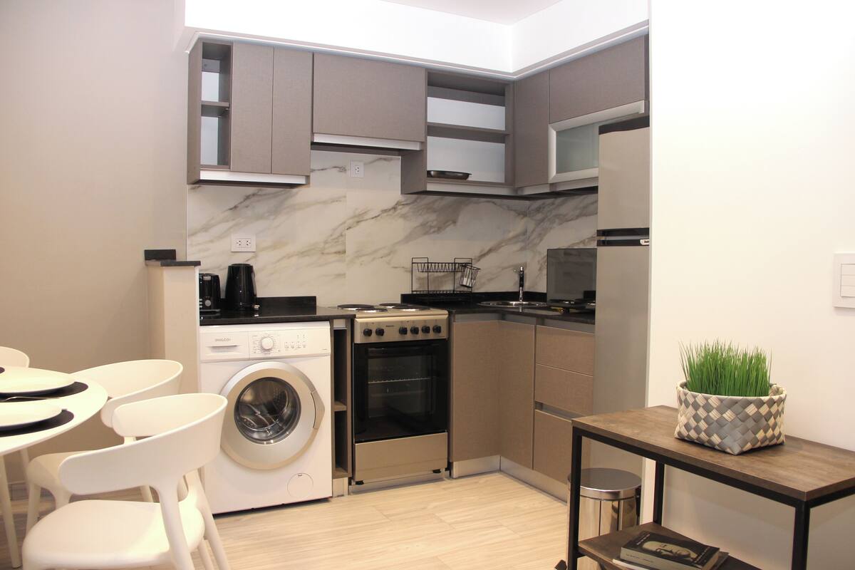 Cocina compacta con lavadora, horno y un pequeño comedor con sillas.