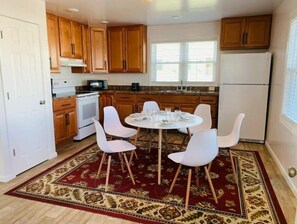 A modern kitchen with wooden cabinets, a white refrigerator, a microwave, and a dining area with white chairs and a red patterned rug.