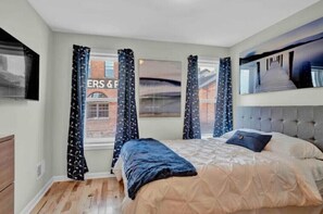Relax in a spacious bedroom with a large bed and tufted headboard, featuring a flat-screen TV and large window with patterned curtains.