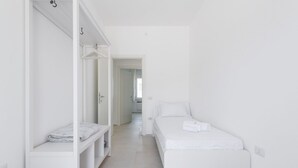 Una habitación minimalista en blanco con una cama individual, un armario empotrado y una puerta visible que da a otra estancia.