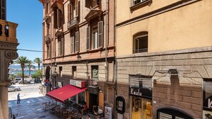 Straßenansicht mit Außenbereich eines Cafés, ein Geschäft mit schwarzem Markisenbalkon und ein Gebäude mit steinverkleideter Fassade.