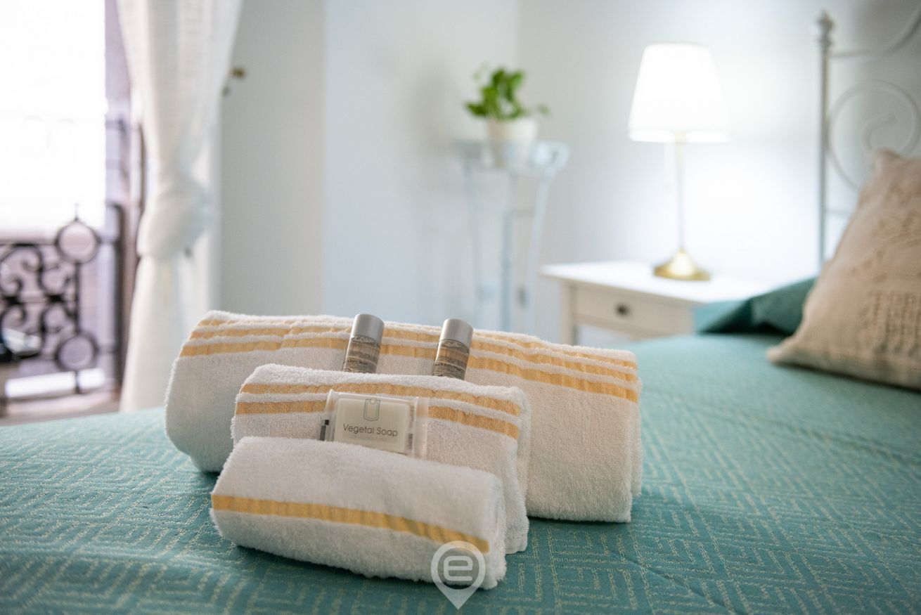 A neatly made bed with a teal bedspread, two rolled towels with a striped pattern, and a small bottle of vegetable soap.