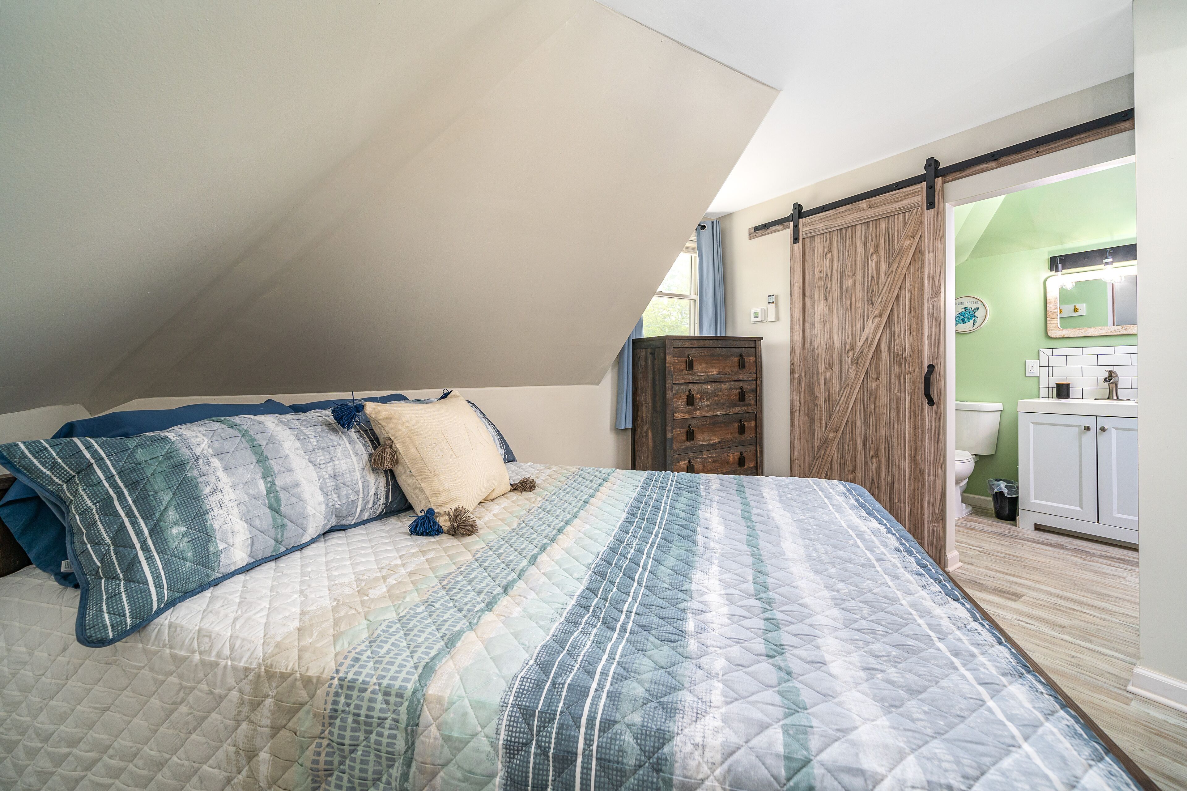 Ein Schlafzimmer mit schrägen Wänden, einem Bett mit gesteppter Bettdecke, einer hölzernen Kommode und einem durchgehenden Türrahmen erkennbaren Badezimmer.