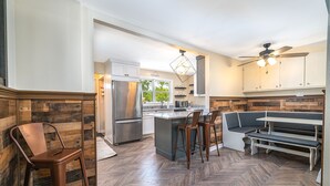 Une cuisine moderne avec un parquet en bois disposé en chevrons, un îlot central intégré avec des tabourets, un coin salon et un ventilateur de plafond.