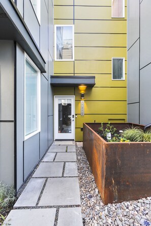 Edifici modern amb un camí estret que condueix a una porta, una jardiniera amb plantes i una petita finestra.