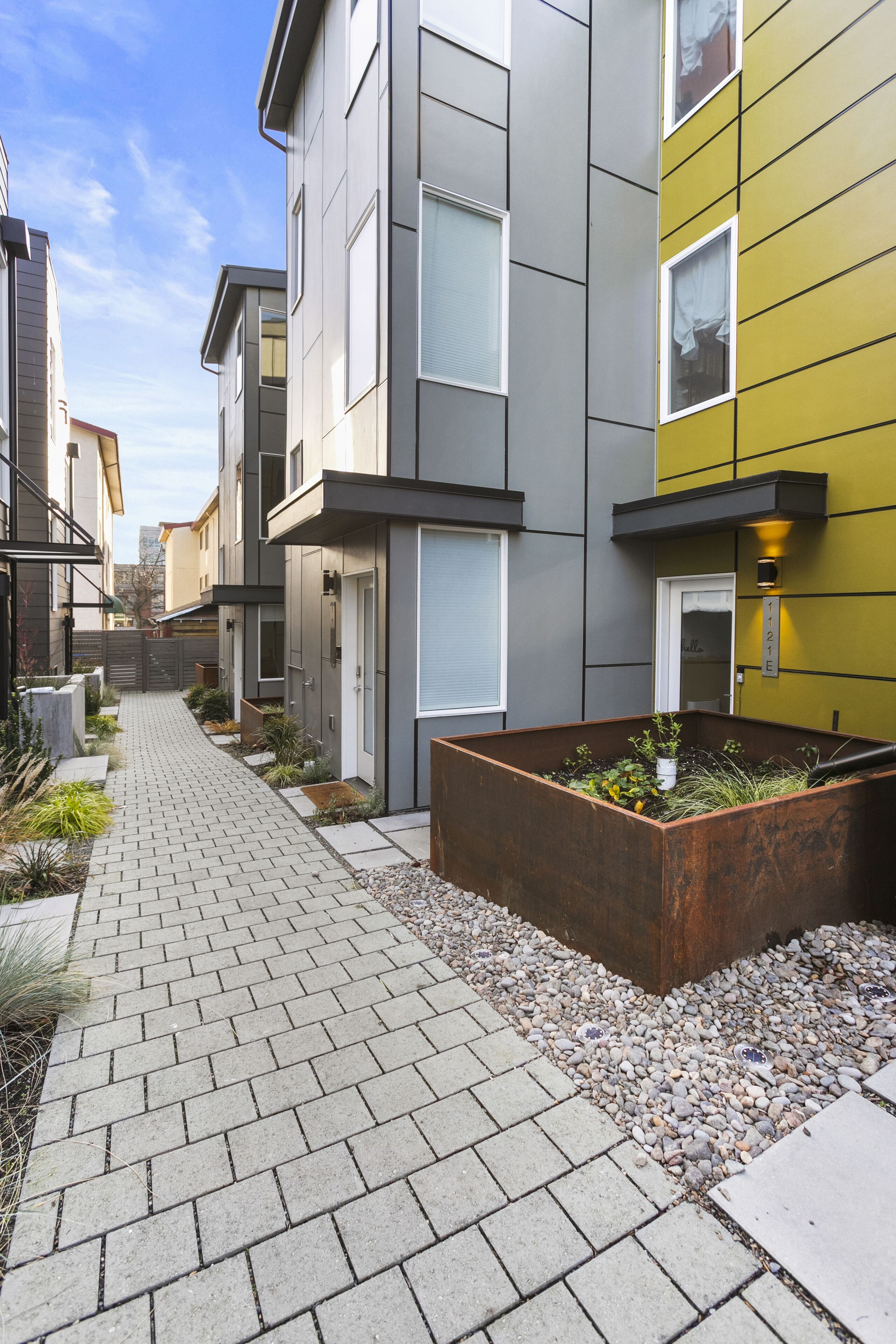 A modern residential building with a paved walkway, a planter box with plants, and a clear sky.