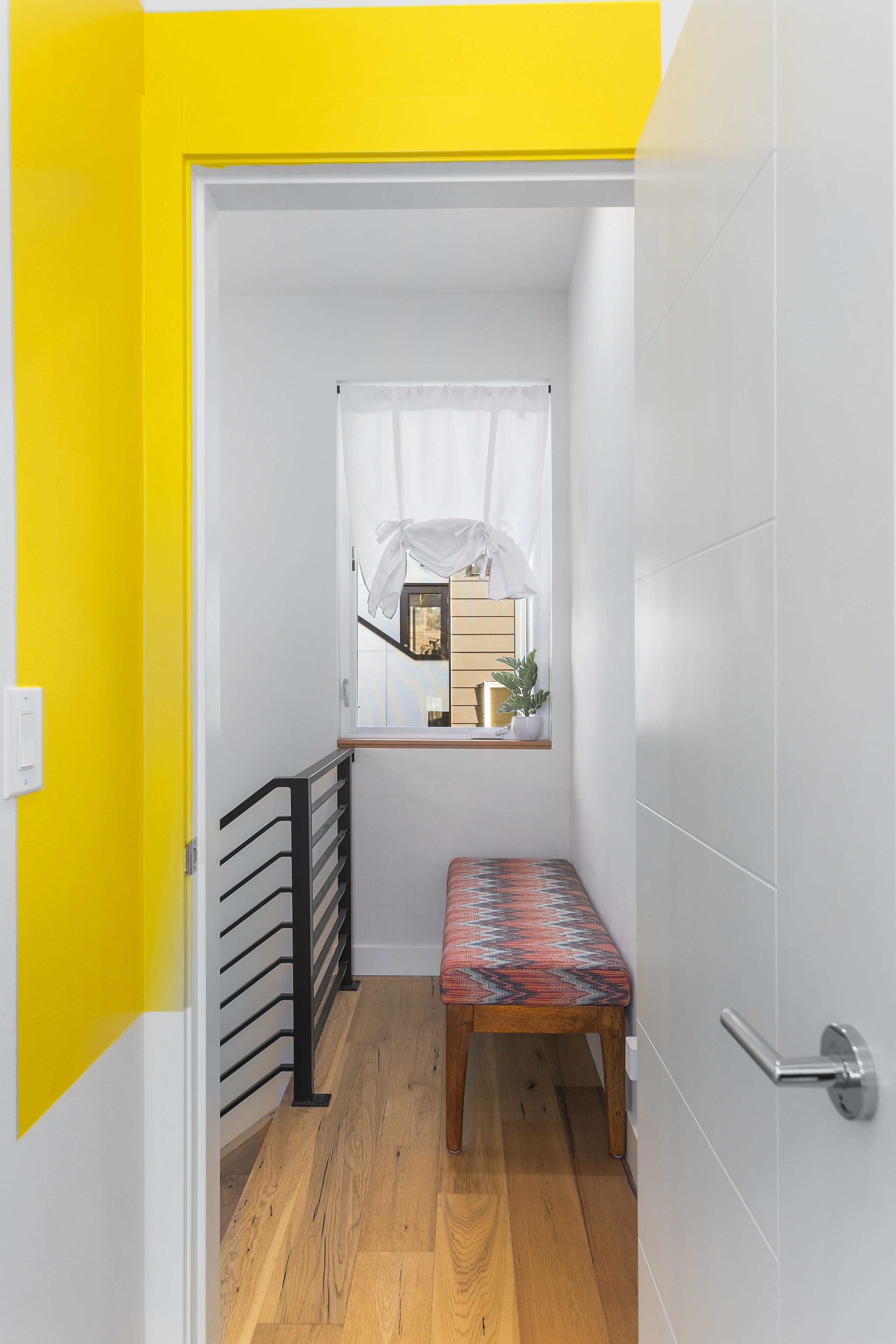 Un pasillo estrecho con un banco, una ventana con cortina y una puerta con marco amarillo.