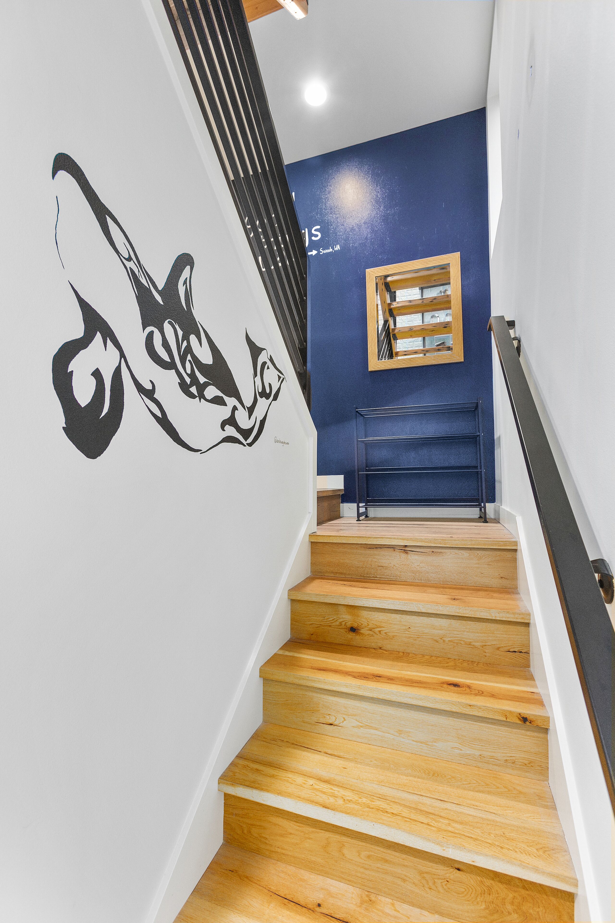 Un escalier en bois, un mur décoré d’un motif noir et blanc, un mur bleu et un petit tableau encadré.