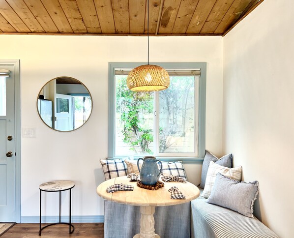 A cozy room with a wooden ceiling, a round mirror, a window with a view of greenery, a round table with a pitcher, and a striped sofa.