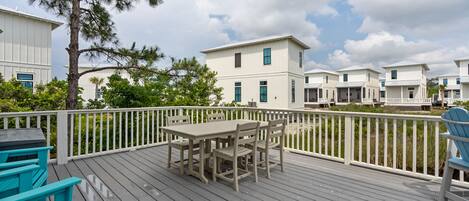 A wooden deck with a table and chairs, surrounded by a white railing, overlooking a row of white buildings.