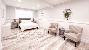 Un dormitorio moderno con cama, dos sillones, una mesita, una mesilla de noche, una ventana y una planta colgante en la pared.