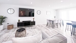 Una sala de estar moderna con televisor de pantalla plana, un comedor con mesa y sillas blancas, y un ventanal con cortinas translúcidas.