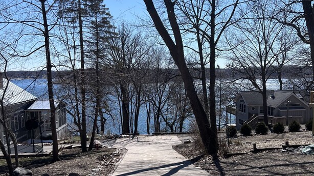 En asfalterad gångväg leder till ett bostadsområde med hus och en sjö eller liknande vattendrag i bakgrunden.