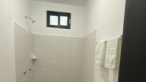 A small, narrow bathroom with white tiles, a toilet, a shower, and a window.