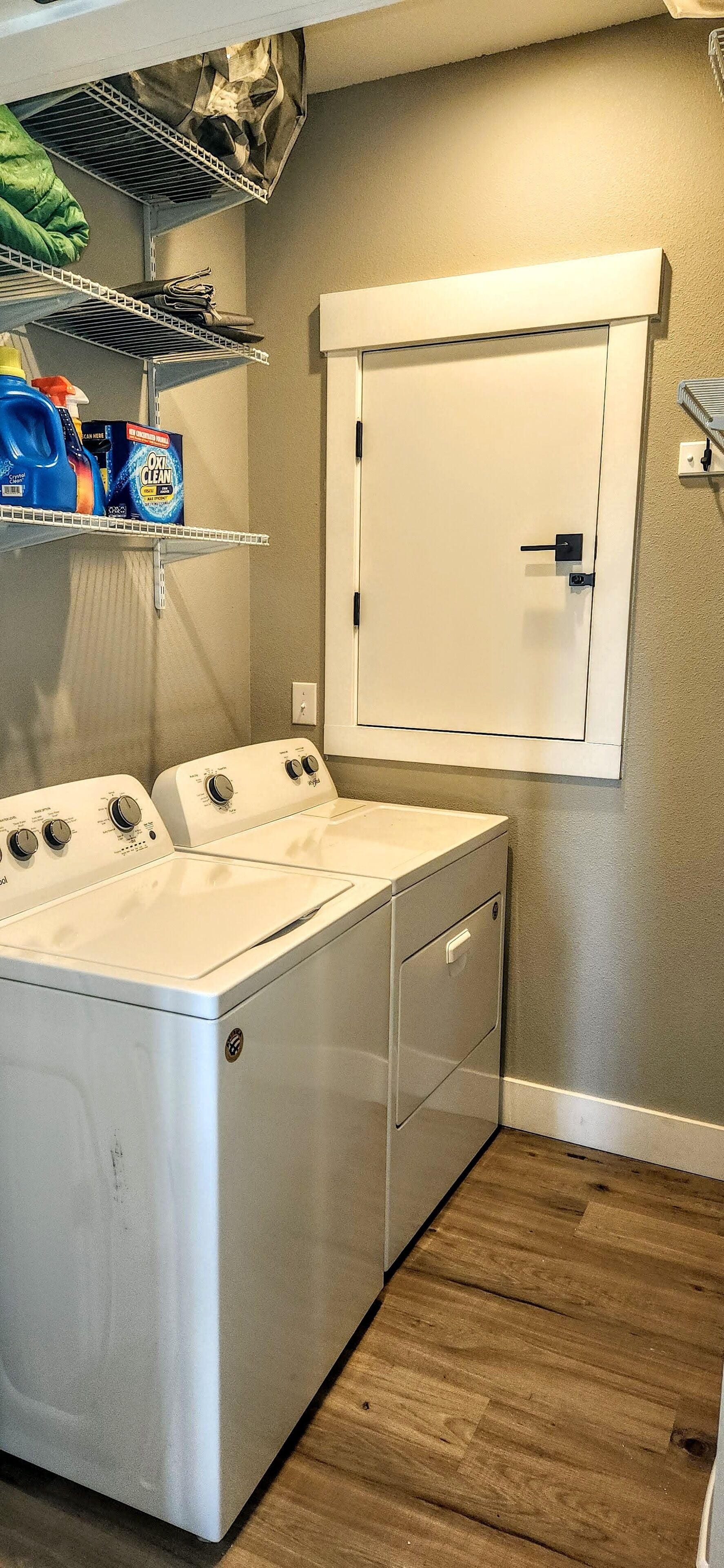 Un cuarto de lavado con dos lavadoras y secadoras apiladas, repisas con productos de limpieza y un armario blanco con puerta.