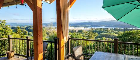 Una terraza de madera con vistas a un lago y un bosque, muebles de exterior y un paraguas verde.