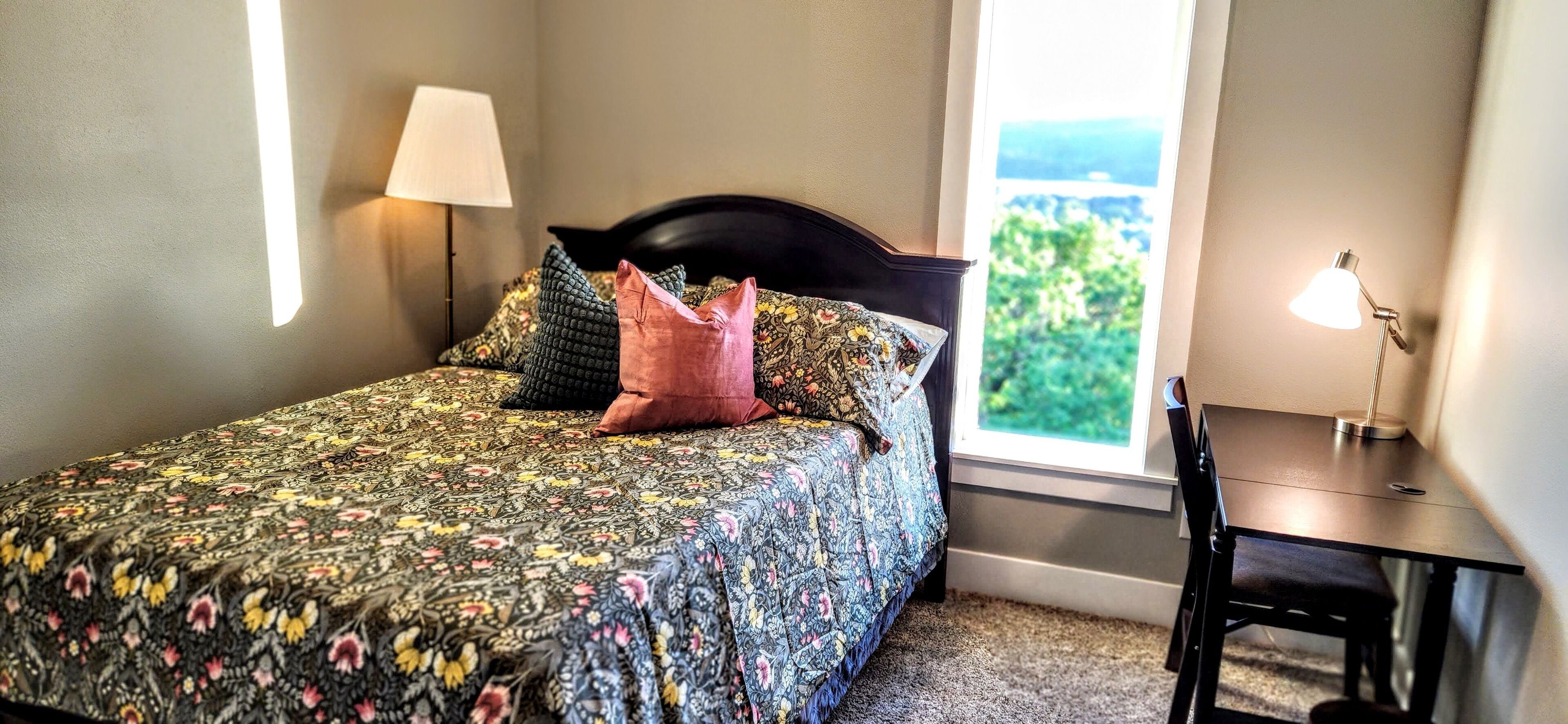 Una cama bien hecha con ropa de cama floral, una mesita de noche con lámpara y vistas a la vegetación a través de una ventana.