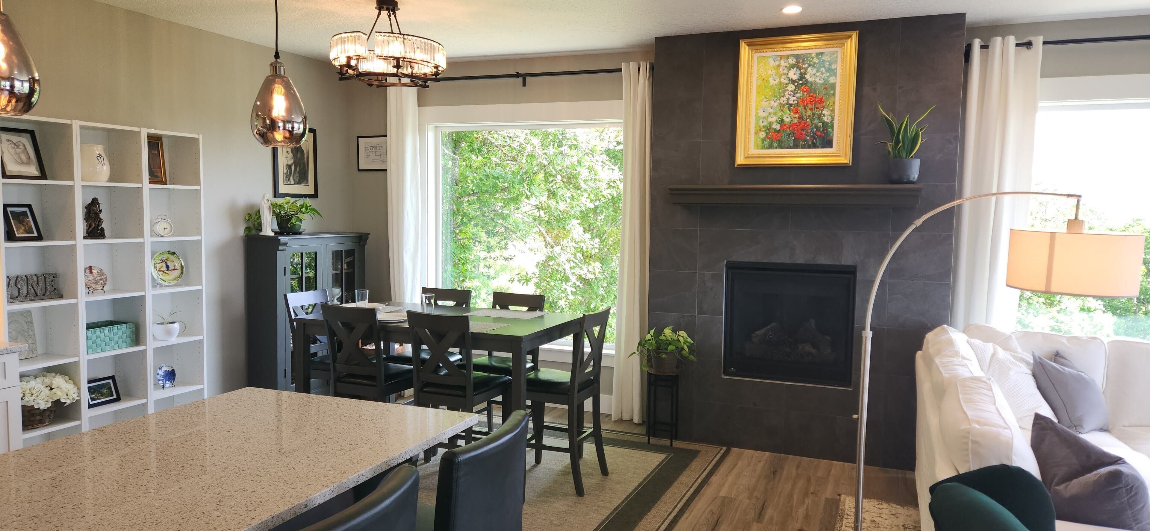 A modern kitchen with a dining area, a fireplace, and a view of greenery outside.