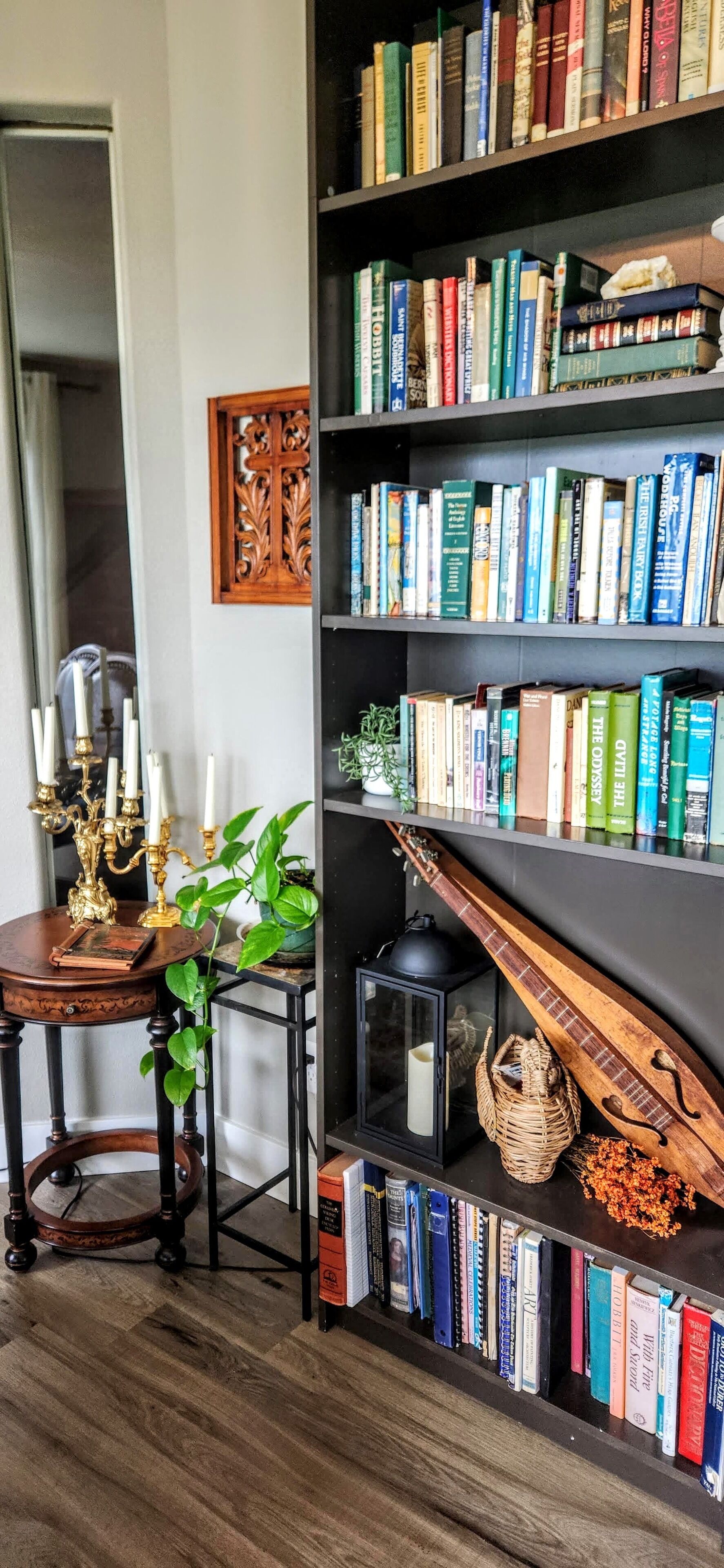 Een kamer met een boekenkast vol boeken, een houten bijzettafeltje met een kandelaar, een kamerplant en een decoratief houten object.