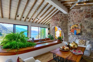 Une pièce spacieuse avec un mur en pierre, des poutres apparentes, une grande fenêtre offrant une vue sur les montagnes et une table en bois agrémentée d’objets décoratifs.