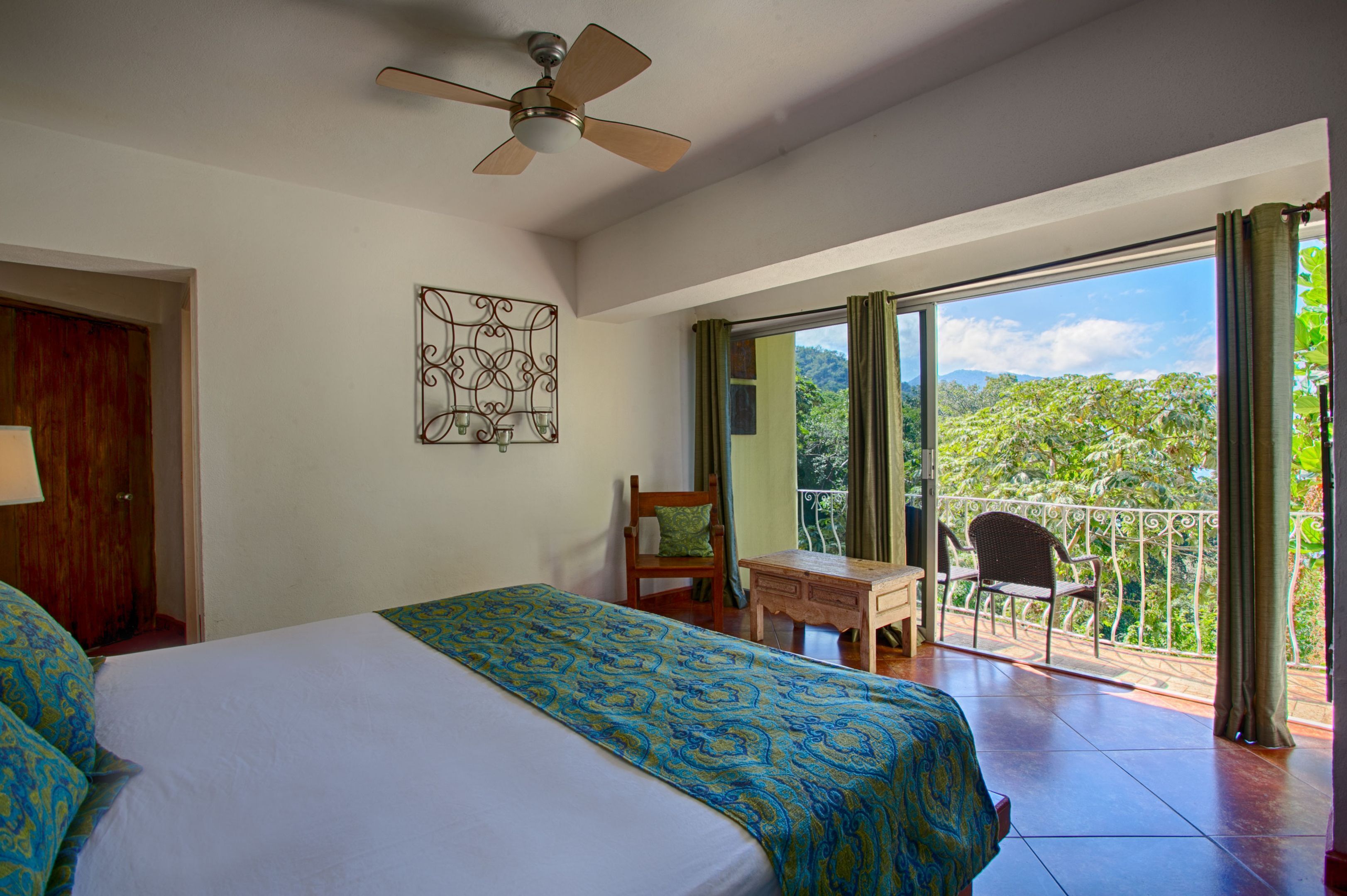 A hotel room with a bed, a desk, and a chair. There is a balcony with a view of greenery.