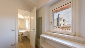 Une pièce avec un plancher en bois, une porte blanche, un bureau blanc avec une chaise, une fenêtre donnant sur des immeubles et une étagère.