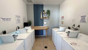 Une buanderie équipée de machines à laver blanches, d’une table en bois et de panneaux indiquant qu’il faut nettoyer le filtre de ventilation et maintenir la propreté des lieux.
