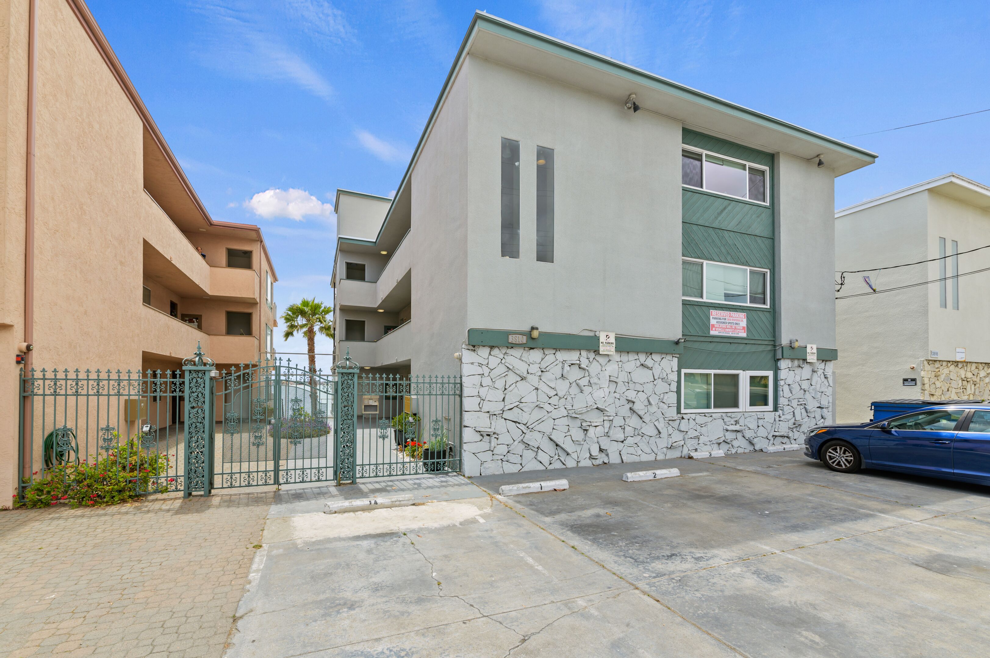 A multi-story residential building with a stone facade, a gated entrance, and a blue car parked in the driveway.