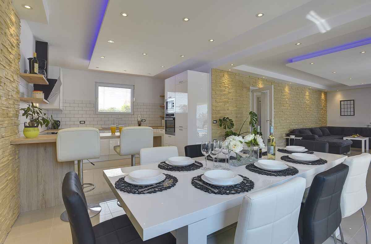 A modern dining area with a long table set for six, white chairs, and a kitchen in the background.