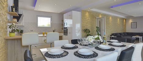 A modern dining area with a long table set for six, white chairs, and a kitchen in the background.