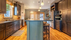Une cuisine moderne avec des meubles de couleur bois foncé, un îlot central, des luminaires suspendus et une vue sur l’extérieur par une fenêtre.